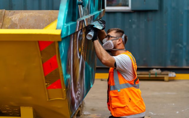 Artist spray painting a skip with vibrant colours as part of the Skip Art Challenge, transforming it into a creative and eye-catching piece of artwork